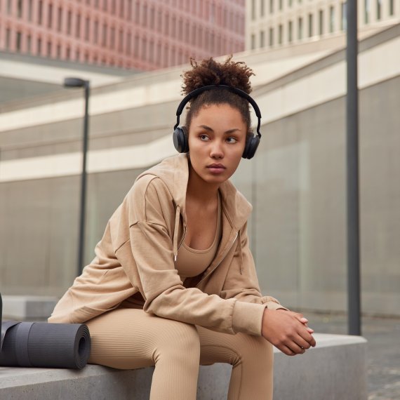 Eine Frau sitzt auf einer Bank in einer Stadt und hört Musik über Kopfhörer. Sie trägt hellbraune bis beige Athleisure Wear.